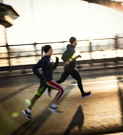 Ein Mann und eine Frau in Sportkleidung laufen bei Sonnenaufgang auf einer sonnigen Brücke für ein Outdoor-Fitnesstraining.