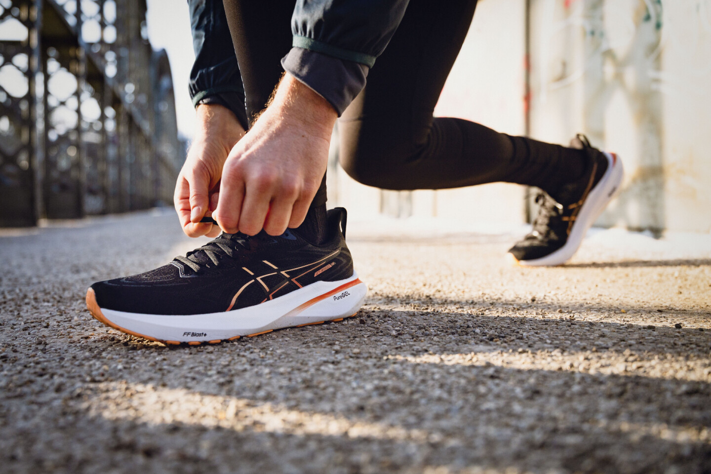 Runner tying black ASICS running shoe laces with orange accents on an urban path.