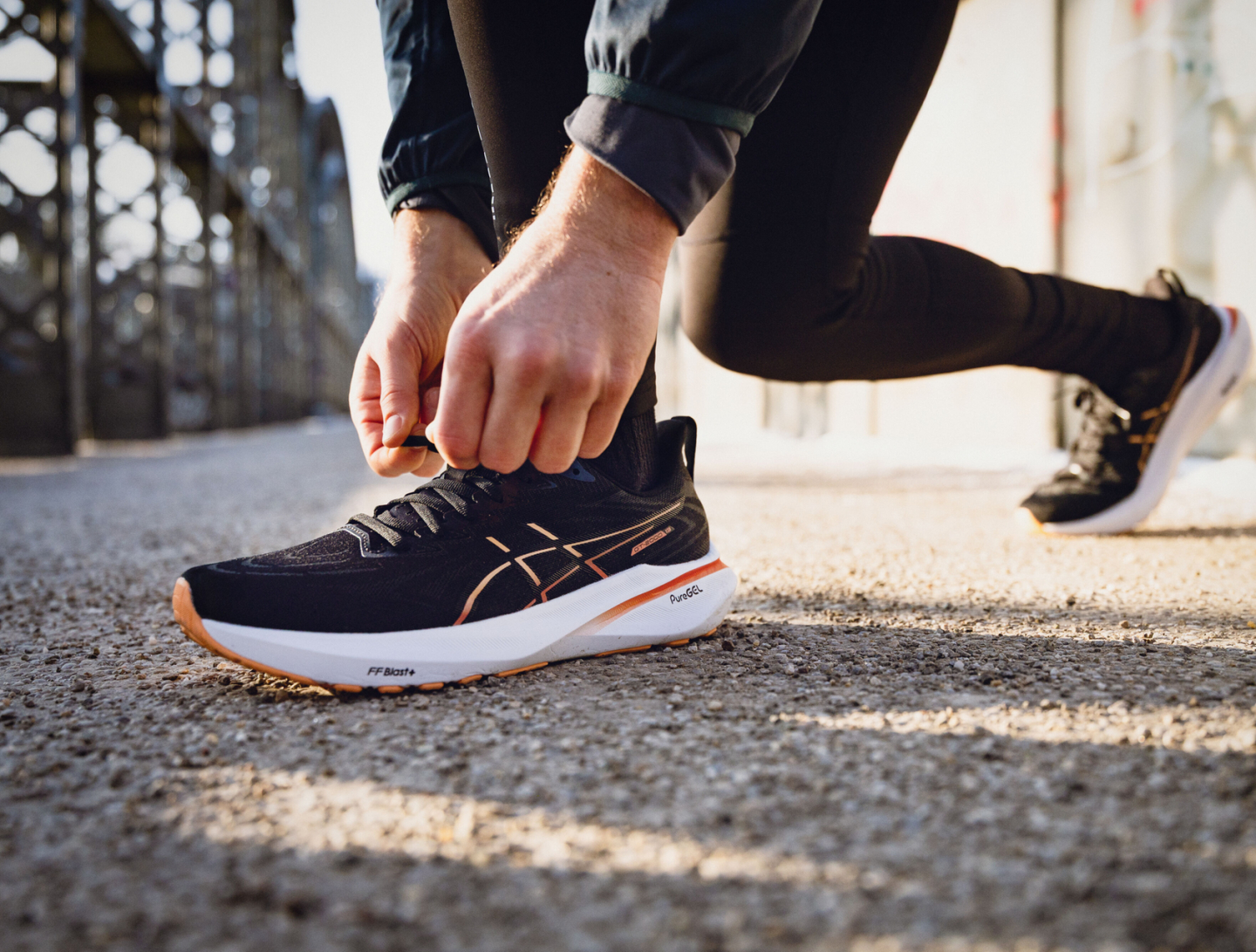 Ein Läufer bindet schwarze ASICS Laufschuhsenkel mit orangefarbenen Akzenten auf einem urbanen Weg.