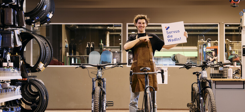 Junger lächelnder Fahrradmechaniker in einer Fahrradwerkstatt, der ein Schild mit der Aufschrift „Servus die Wadln!“ in der Nähe von Fahrrädern hält.
