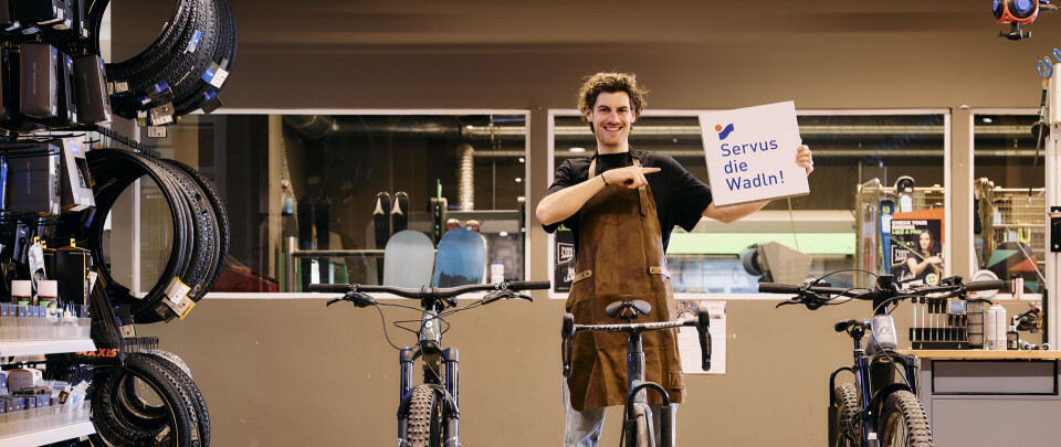 Junger, lächelnder Fahrradmechaniker in einer Fahrradwerkstatt, der ein Schild mit der Aufschrift „Service de Wielen!“ in der Nähe von Fahrrädern hält.