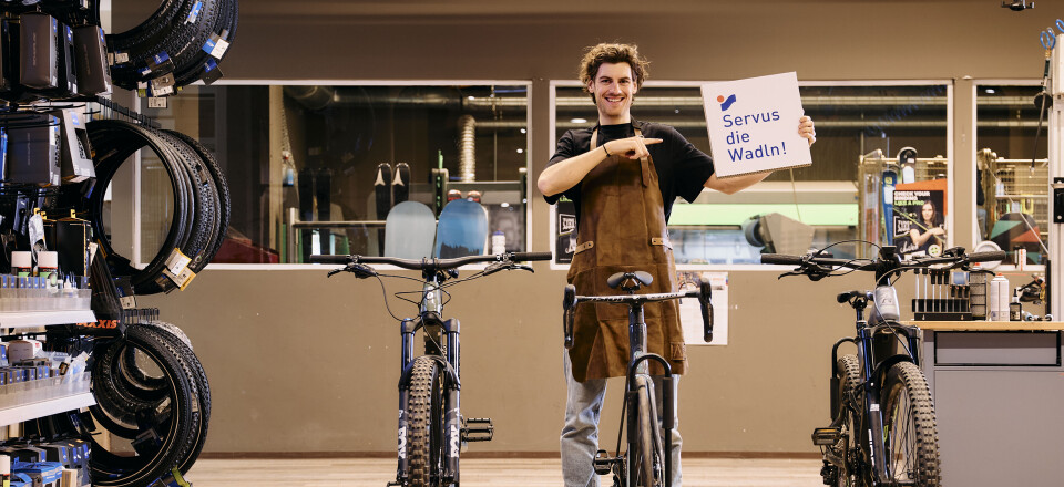 Junger lächelnder Fahrradmechaniker in einer Fahrradwerkstatt, der ein "Servus die Wadln!"-Schild neben Fahrrädern hält.