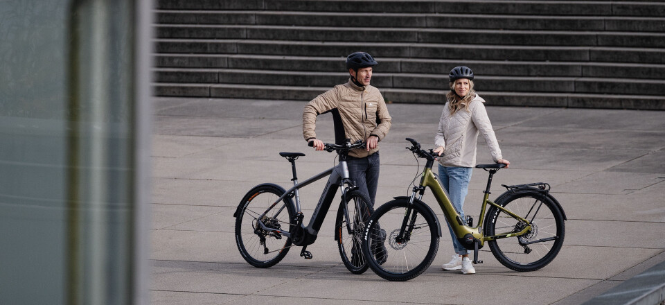 Ein Paar mit Helmen steht mit zwei E-Bikes auf einem modernen Stadtplatz, im Hintergrund Betontreppen.