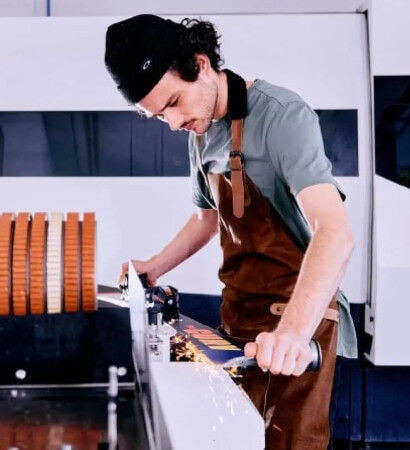 Ein Mann in Schürze präpariert einen Ski an einer modernen Maschine in einer Werkstatt, mit einer großen Walze orangefarbener Scheiben.