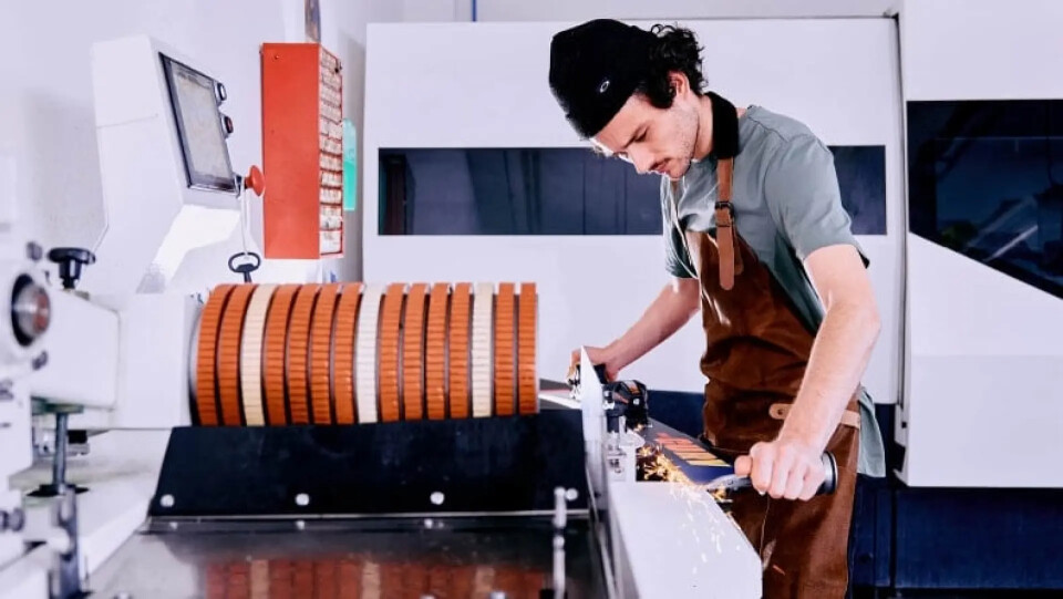 Ein Mann in Schürze präpariert einen Ski an einer modernen Maschine in einer Werkstatt, mit einer großen Walze orangefarbener Scheiben.