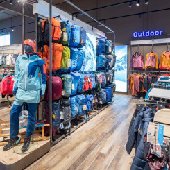 Innenansicht eines Outdoor-Einzelhandelsgeschäfts mit einer Schaufensterpuppe in blauer Jacke, leuchtenden Rucksäcken und Kleiderauslagen.