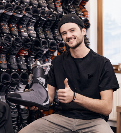 Lächelnder Mann in einem Skiverleih hält einen Skischuh, zeigt den Daumen nach oben, umgeben von Wintersportausrüstung.