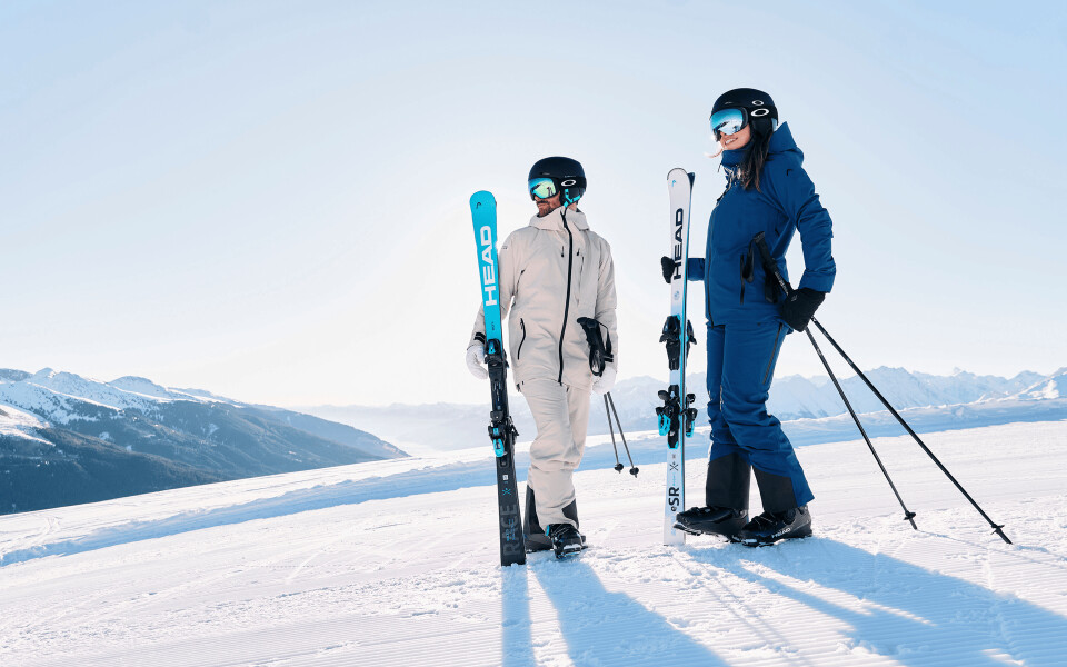 Zwei Skifahrer in blauen und beigefarbenen Skianzügen, Helmen und Skibrillen stehen mit Skiern und Skistöcken auf einem sonnigen Berghang.
