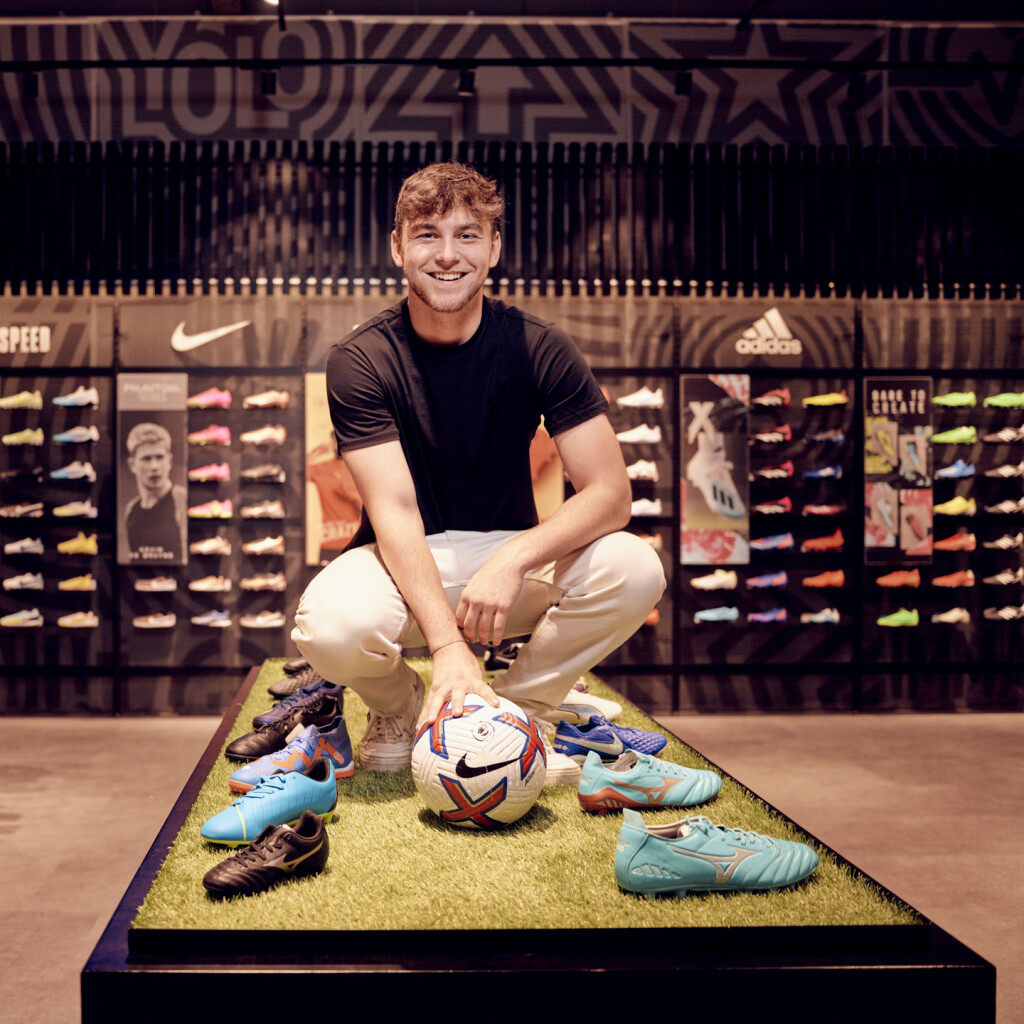 Ein lächelnder Mann hockt mit einem Fußball und bunten Sportschuhen in einem Sportfachgeschäft.