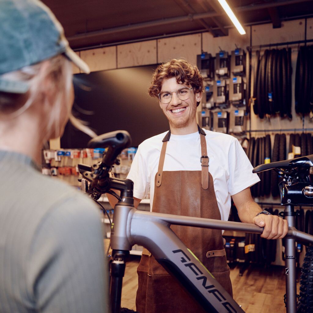 Ein lächelnder Fahrradladenmitarbeiter mit Schürze hilft einer Kundin bei einem neuen E-Bike in einem modernen Fahrradgeschäft.