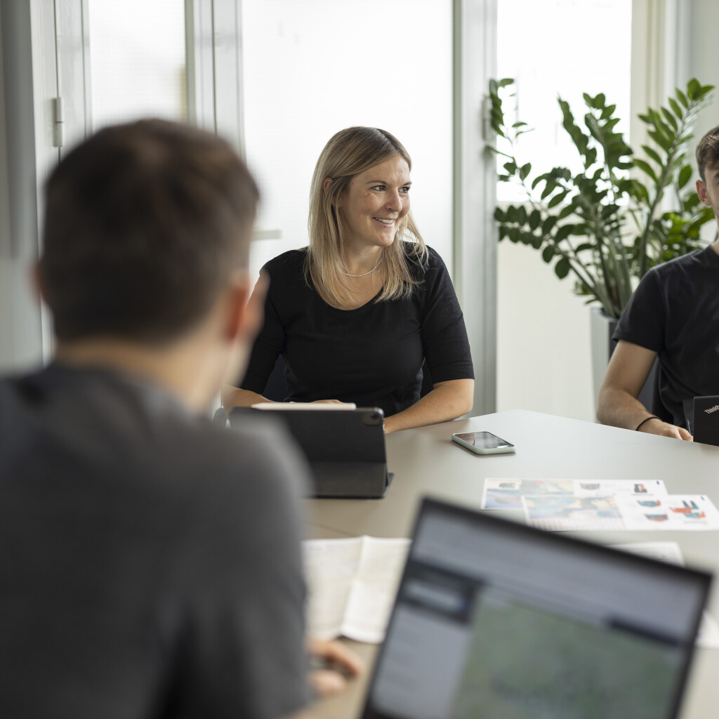 Geschäftsleute in einem Büromeeting, die an einem Tisch zusammenarbeiten, mit Laptops und einem großen Bildschirm, der eine Karte zeigt.