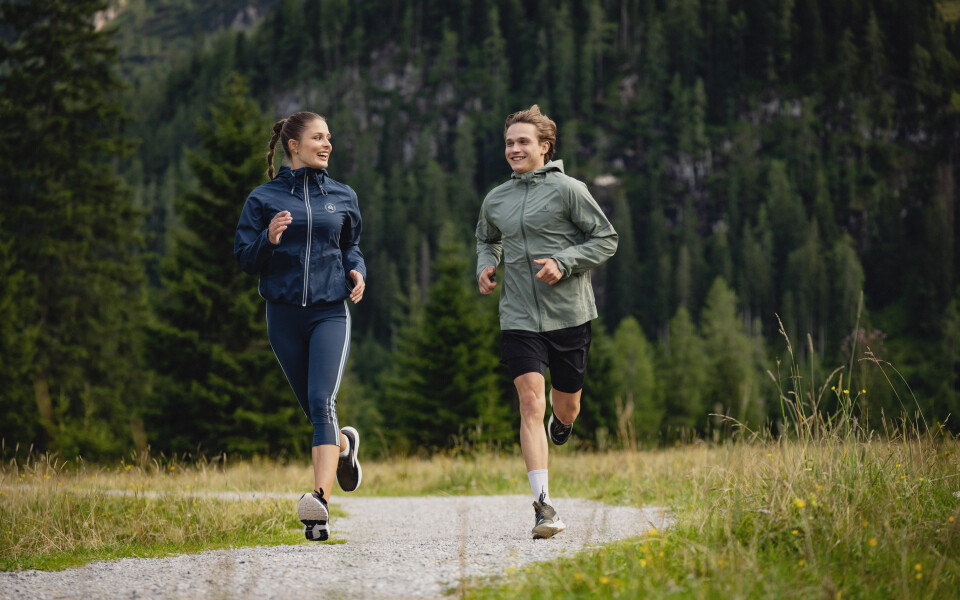 Zwei lächelnde Personen beim Trailrunning auf einem Schotterweg durch einen leuchtend grünen Wald mit Bergen.