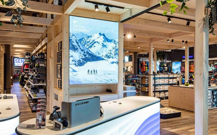 Modernes Ladeninterieur mit einer beleuchteten wellenförmigen Theke, Holzbalken, Hängepflanzen und einem großen digitalen Bergdisplay.