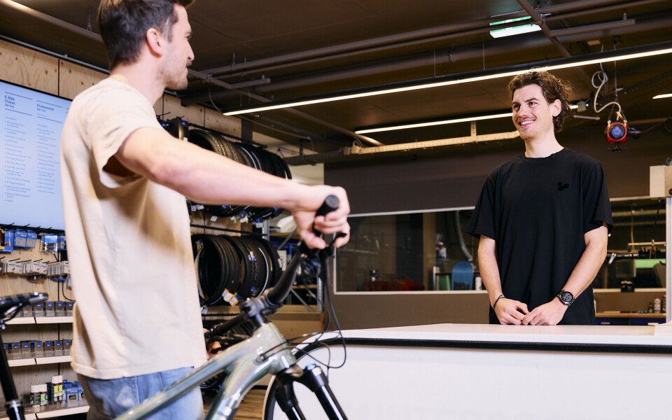 Ein Kunde mit einem Mountainbike spricht mit einem lächelnden Mitarbeiter an der Theke eines modernen Fahrradgeschäfts.